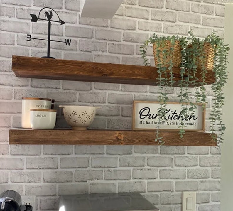May include: Two rustic, stained wooden shelves mounted on a brick-patterned wall. The top shelf holds two woven baskets with trailing greenery. The bottom shelf displays a sign that reads "Our Kitchen" and two ceramic containers.