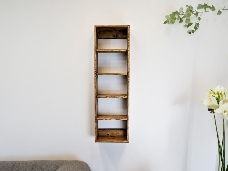 May include: A tall, narrow wooden shelf with six open compartments. The shelf is made of stained wood, showcasing the natural grain and knots. It is mounted on a white wall, with a simple, rustic design. The shelf is ideal for displaying books or decorative items.