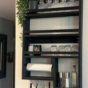 May include: A black wooden kitchen shelf unit mounted on a wall, featuring multiple shelves for storage. It holds champagne flutes, glassware, a paper towel roll, spices, and utensils. A potted plant and decorative items are also present.