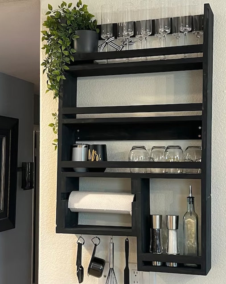 May include: A black wooden kitchen shelf unit mounted on a wall, featuring multiple shelves for storage. It holds champagne flutes, glassware, a paper towel roll, spices, and utensils. A potted plant and decorative items are also present.