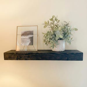 May include: A dark stained wooden floating shelf with a framed black and white photograph and a decorative vase with green foliage. The shelf is mounted on a cream-colored wall, creating a minimalist aesthetic.