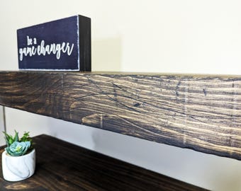 May include: A dark stained wooden shelf with a decorative block sign that reads "be a game changer" in white script. A small potted succulent sits on a lower shelf. The wood grain is visible.