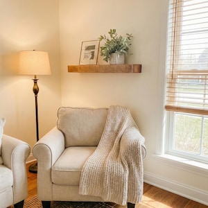 May include: A cozy living room scene featuring a beige armchair with a knitted throw blanket. A wooden shelf displays a framed picture and potted greenery. A floor lamp and a window with blinds complete the warm, inviting space.