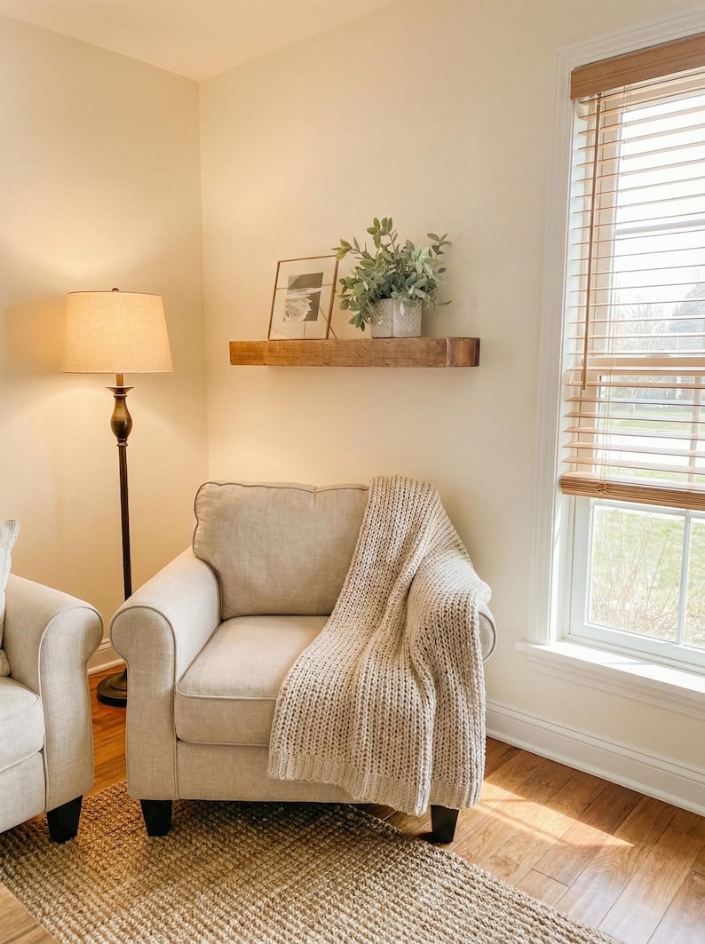 May include: A cozy living room scene featuring a beige armchair with a knitted throw blanket. A wooden shelf displays a framed picture and potted greenery. A floor lamp and a window with blinds complete the warm, inviting space.