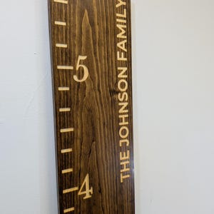 May include: A wooden height chart with engraved markings and the text "THE JOHNSON FAMILY." The chart is stained dark brown with visible wood grain. Height markers are in feet, with the numbers 4 and 5 visible.
