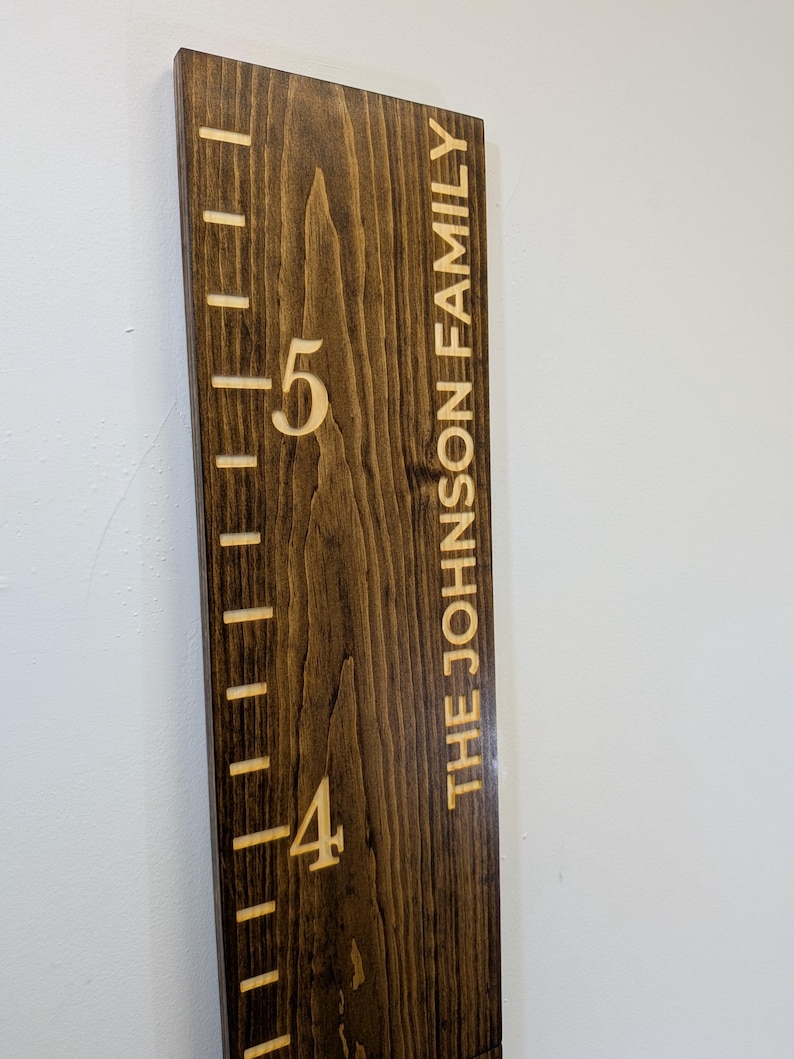 May include: A wooden height chart with engraved markings and the text "THE JOHNSON FAMILY." The chart is stained dark brown with visible wood grain. Height markers are in feet, with the numbers 4 and 5 visible.