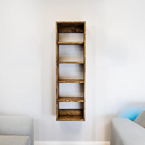 May include: A tall, narrow, wooden bookshelf with six shelves, mounted on a white wall. The shelves are stained a warm brown color, and the wood grain is visible. The bookshelf is empty, ready for display.