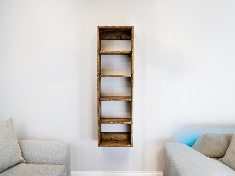 May include: A tall, narrow, wooden bookshelf with six shelves, mounted on a white wall. The shelves are stained a warm brown color, and the wood grain is visible. The bookshelf is empty, ready for display.