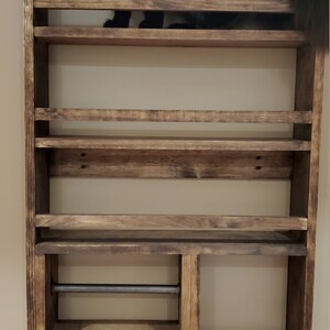 May include: A rustic, wooden spice rack with multiple shelves, featuring a built-in paper towel holder. A black cat is perched on the top shelf, adding a playful element to the design. The rack is mounted on a neutral-colored wall.