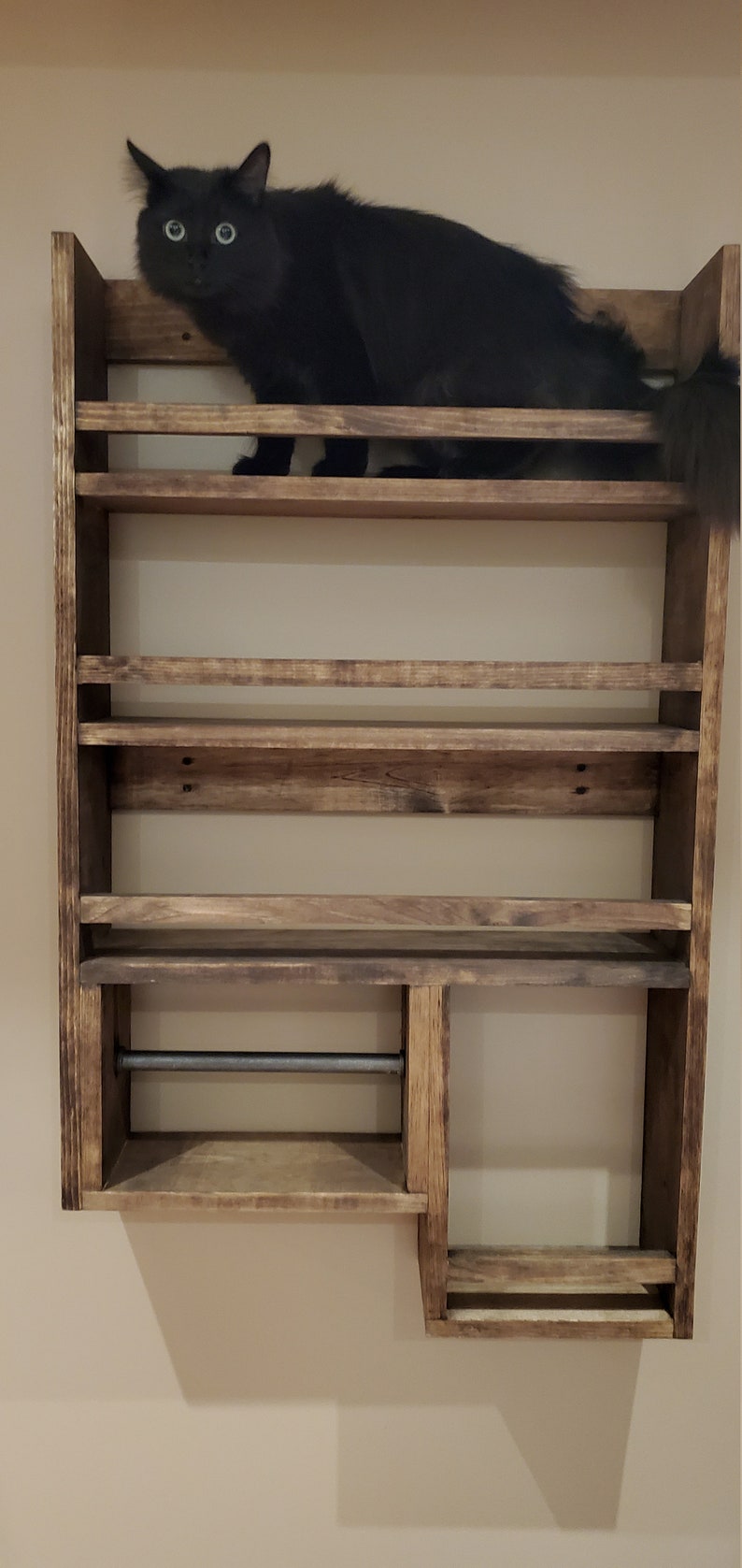 May include: A rustic, wooden spice rack with multiple shelves, featuring a built-in paper towel holder. A black cat is perched on the top shelf, adding a playful element to the design. The rack is mounted on a neutral-colored wall.