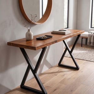 Peut inclure: Une console rectangulaire en bois avec une finition marron naturel et une surface lisse. La table dispose de deux pieds en métal noir en forme de X. Sur la table se trouvent un petit vase en céramique beige avec des tiges, un plateau noir et des livres.