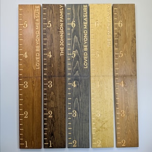May include: Wooden height charts with engraved measurements and family names. The charts are stained in various shades of brown, grey, and gold. Each chart displays the text "LOVED BEYOND MEASURE" or a family name, with measurements in feet.