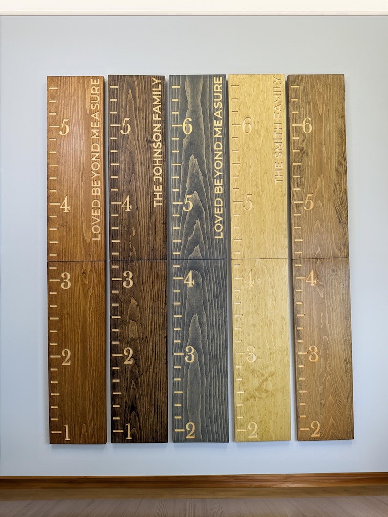 May include: Wooden height charts with engraved measurements and family names. The charts are stained in various shades of brown, grey, and gold. Each chart displays the text "LOVED BEYOND MEASURE" or a family name, with measurements in feet.