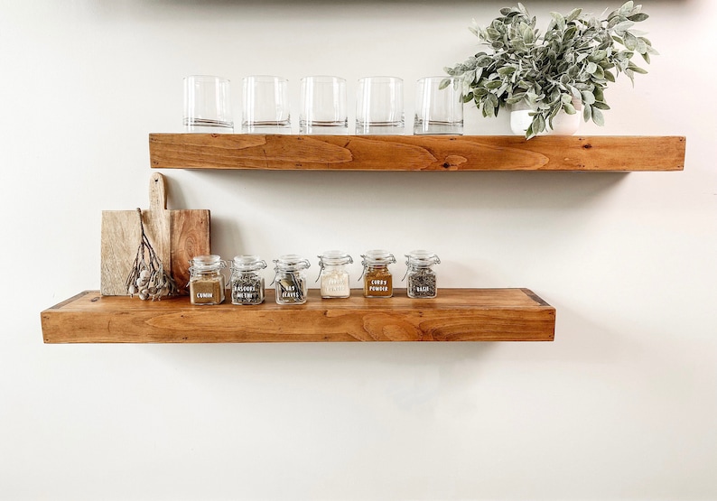 K&ouml;nnte beinhalten: Zwei Holzregale an einer wei&szlig;en Wand. Das obere Regal enth&auml;lt sechs klare Trinkgl&auml;ser und eine Topfpflanze. Das untere Regal zeigt sechs kleine Gl&auml;ser mit Etiketten und ein Holzschneidebrett.