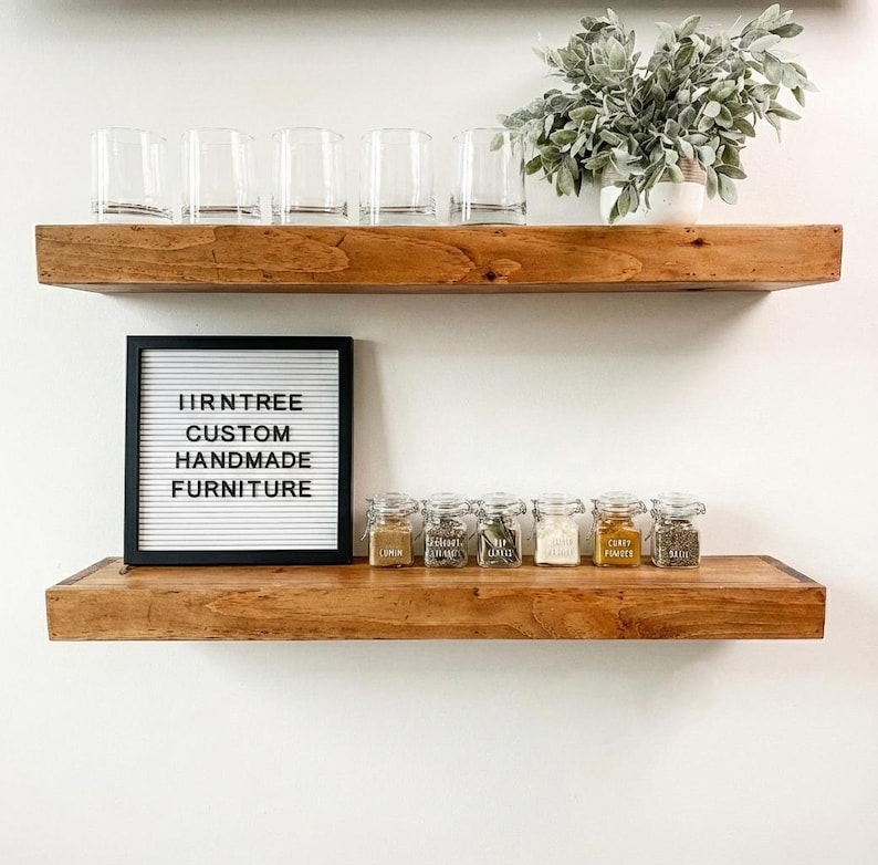 May include: Two wooden shelves with various items. The top shelf holds six clear glasses and a potted plant. The bottom shelf displays a letter board with text and small glass jars of spices. The letter board reads "HRNTREE CUSTOM HANDMADE FURNITURE."