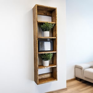 May include: A tall, rustic wooden shelf unit with four shelves, mounted on a white wall. The shelves hold two potted plants and a framed picture. The wood has a warm, natural finish, and the design is simple and modern.