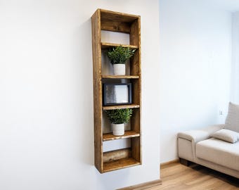 May include: A tall, rustic wooden shelf unit with four shelves, mounted on a white wall. The shelves hold two potted plants and a framed picture. The wood has a warm, natural finish, and the design is simple and modern.