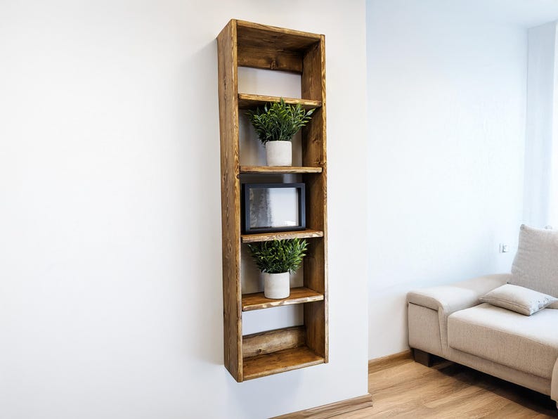 May include: A tall, rustic wooden shelf unit with four shelves, mounted on a white wall. The shelves hold two potted plants and a framed picture. The wood has a warm, natural finish, and the design is simple and modern.