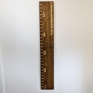May include: A dark stained wooden height chart with etched markings from 1 to 5 feet. The text "THE JOHNSON FAMILY" is vertically etched on the right side. The chart is mounted on a white wall.
