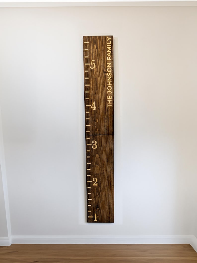 May include: A dark stained wooden height chart with etched markings from 1 to 5 feet. The text "THE JOHNSON FAMILY" is vertically etched on the right side. The chart is mounted on a white wall.