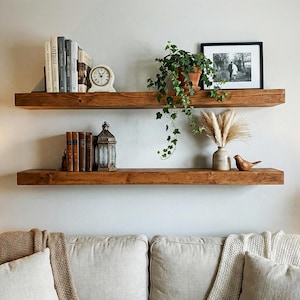 May include: Two rustic, wooden floating shelves mounted on a light-colored wall. The top shelf holds books, a clock, and a potted plant. The bottom shelf displays books, a lantern, a vase with dried flowers, and a wooden bird figurine. A sofa is below.
