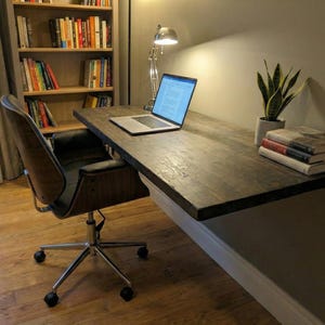May include: A dark wood desk is mounted to the wall, with a laptop and books on top. A black leather office chair with a chrome base sits in front of the desk. A bookshelf and a potted plant are also visible.