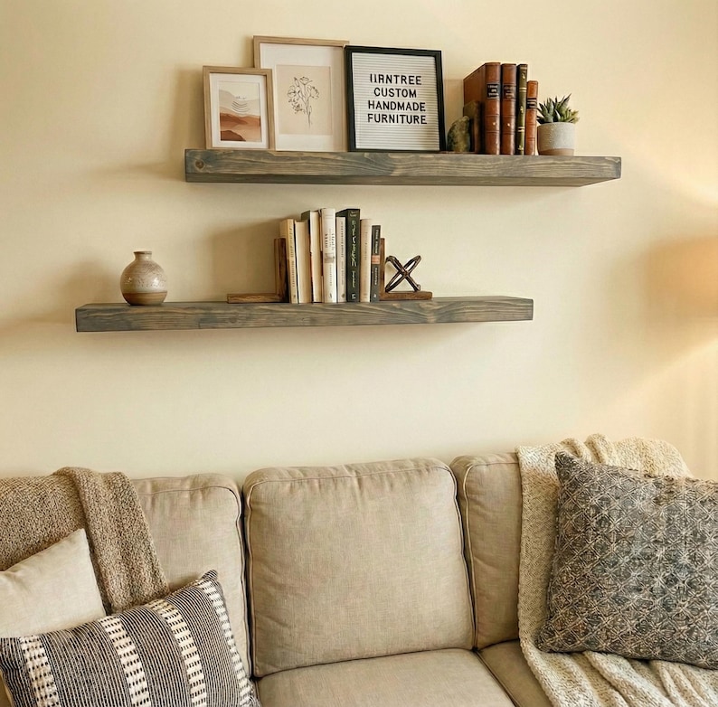 May include: Two gray wooden floating shelves mounted on a cream-colored wall. The top shelf holds framed art, books, and a small plant. The bottom shelf displays books and a decorative vase. The text on the sign reads "IIRNTREE CUSTOM HANDMADE FURNITURE."