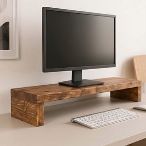 May include: A rectangular monitor stand crafted from dark stained wood with a visible grain pattern. The stand features two sturdy side supports and a flat top surface, currently holding a black computer monitor on a desk.