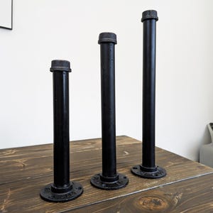 May include: Three black metal pipe flanges with different lengths. The flanges are on a wooden surface.