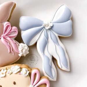 Puede incluir: Una galleta en forma de lazo azul y blanco con una flor blanca en la parte superior. La galleta está decorada con glaseado real.