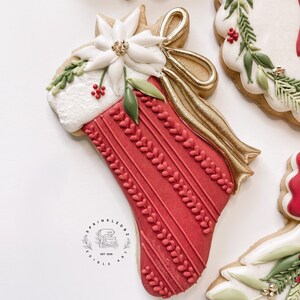 Puede incluir: Una galleta de calcetín de Navidad roja y blanca decorada con una flor de pascua blanca, hojas verdes y una cinta dorada. La galleta está decorada con un patrón de rayas rojas y blancas y tiene un pequeño logotipo en la parte inferior que dice "Sprinkledbz Edible Art Est. 2020".