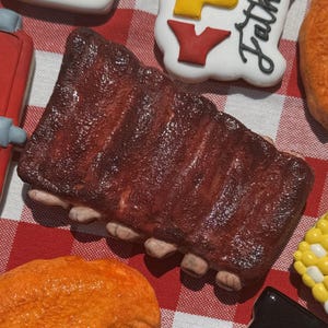May include: A set of nine decorated sugar cookies with a barbecue theme. The cookies are shaped like a beer mug, ribs, chicken wings, corn on the cob, a bottle of barbecue sauce, and a sign that says "Happy Father's Day".