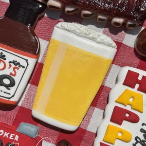 May include: A set of five decorated sugar cookies on a red and white checkered tablecloth. The cookies are shaped like a beer glass, a bottle of barbecue sauce, a grill, ribs, and a sign that says "Happy Father's Day".