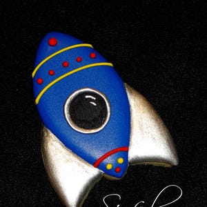 May include: A blue rocket-shaped cookie with yellow and red accents, and silver wings. The cookie has a circular window and is decorated with red dots and yellow stripes. The text "Sinful Decadence" is written in cursive.