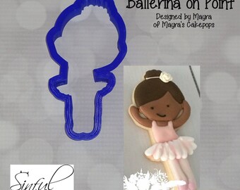 Op de afbeelding: Blauwe plastic koekjesvorm in de vorm van een ballerina. De afbeelding toont ook een versierde koek met een ballerina-ontwerp, met een roze tutu. Tekst luidt "Ballerina on Point" en "Sinful Cutters".
