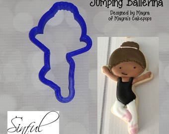 Op de afbeelding: Een blauwe plastic koekjesvorm in de vorm van een ballerina, naast een versierde koek. De koek is een ballerina met een zwart turnpakje, witte panty en roze balletschoenen. De tekst "Jumping Ballerina" en "Sinful Cutters" staan op de afbeelding.