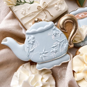 Op de afbeelding: Een lichtblauwe theepotvormige koek met witte bloemendetails en gouden rand. Andere koekjes zijn een rechthoekige met een strik en een ronde met een gouden handvat. De koekjes zijn versierd met witte glazuur.