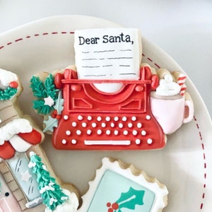 May include: A red and white gingerbread cookie shaped like a vintage typewriter with the words "Dear Santa" on a piece of paper in the typewriter. The cookie is on a white plate with other gingerbread cookies.