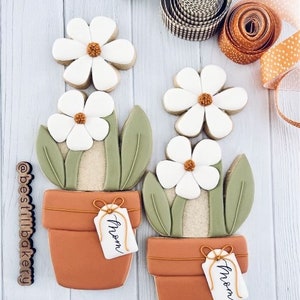 May include: Two decorated sugar cookies shaped like potted plants with white flowers and green leaves. Each pot has a brown tag that says "Mom".