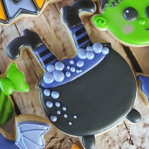May include: A black and purple witch's cauldron cookie decorated with purple and white icing dots. The witch's legs are sticking out of the top of the cauldron.