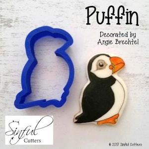 May include: A blue plastic puffin-shaped cookie cutter next to a decorated puffin cookie. The cookie has a black body, white belly, orange beak and feet, and a detailed eye. Text on the image reads "Puffin" and "Decorated by Angie Brechtel."