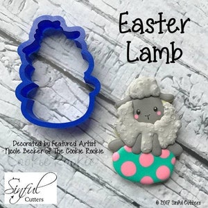 May include: Blue plastic cookie cutter shaped like a lamb sitting on an egg. The egg is decorated with pink and teal polka dots. The cookie cutter is next to a decorated sugar cookie of a lamb sitting on an egg. The cookie is decorated with white frosting and has a gray face with black eyes and a pink nose. The text "Easter Lamb" is written in black above the cookie.