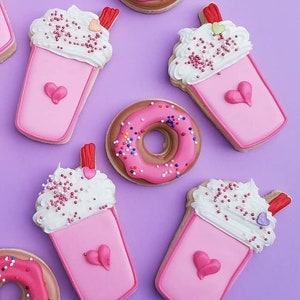 May include: An assortment of decorated sugar cookies in the shape of pink milkshakes and donuts. The milkshake cookies have white frosting, sprinkles, and a heart design. The donuts are pink with sprinkles.