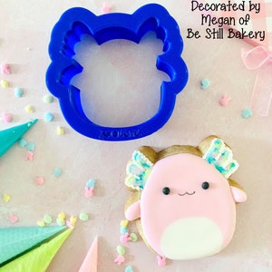 May include: A blue plastic axolotl cookie cutter next to a decorated axolotl cookie. The cookie is pink with a white belly, brown accents, and colorful details. Text on the image reads "Axolotl Fluffy Fellow" and "Decorated by Megan of Be Still Bakery."