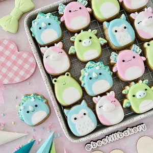 Puede incluir: Primer plano de una bandeja para hornear con una variedad de galletas de azúcar decoradas. Las galletas tienen forma de animales, incluyendo un unicornio, un dinosaurio, una rana, un búho y un dragón. Las galletas están decoradas con glaseado de colores y chispas. La bandeja para hornear está sobre una superficie rosa con un corazón a cuadros rosa y blanco.