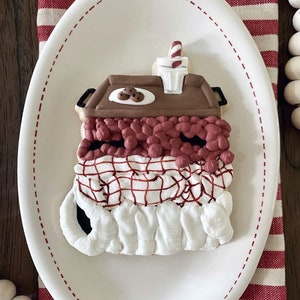 Può includere: Un piatto in ceramica bianca con un tovagliolo a righe rosse e bianche sotto. Sul piatto c'è un biscotto di zucchero decorato a forma di calza di Natale riempita di bastoncini di zucchero rossi e bianchi e un piccolo cartone di latte con una cannuccia a righe rosse e bianche.
