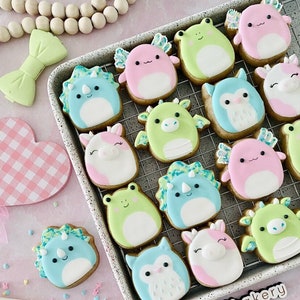 May include: A selection of decorated sugar cookies on a wire rack. The cookies are decorated with various animal designs, including a unicorn, a dinosaur, a dragon, an owl, and a frog. The cookies are all in pastel colours, and the rack is on a pink surface. The text "@bestillbakery" is visible on the surface.