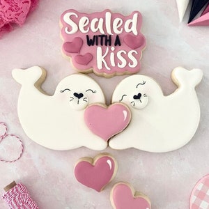 Op de afbeelding: Twee witte suikerkoekjes versierd als zeehonden die een roze hartkoekje vasthouden. De koekjes liggen op een roze achtergrond met een roze en wit geruit koekje en een roze koekje met de tekst "Sealed with a Kiss" in zwart.