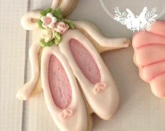 Op de afbeelding: Een paar ballet-schoen koekjes met roze binnenkant en lichtroze buitenkant. De koekjes zijn versierd met kleine roze en witte bloemen en groene blaadjes. De koekjes liggen op een lichtgekleurd oppervlak.