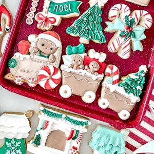May include: A set of Christmas-themed sugar cookies decorated with icing. The cookies feature a train, a fireplace, a Christmas tree, and a candy cane. The cookies are on a red baking sheet with a white and red striped background.
